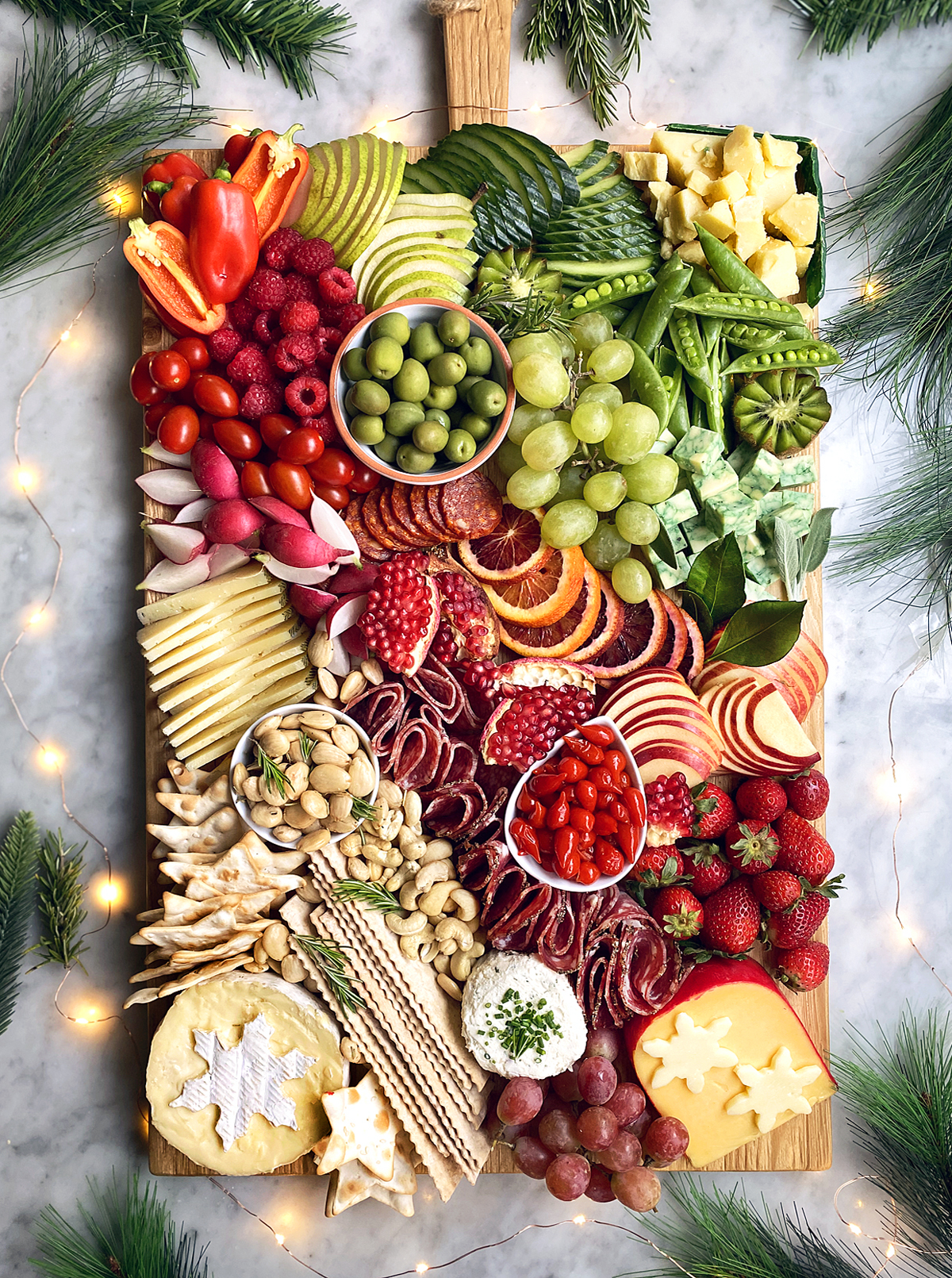 red, green, white cheese and charcuterie on wooden board with christmas lights in background