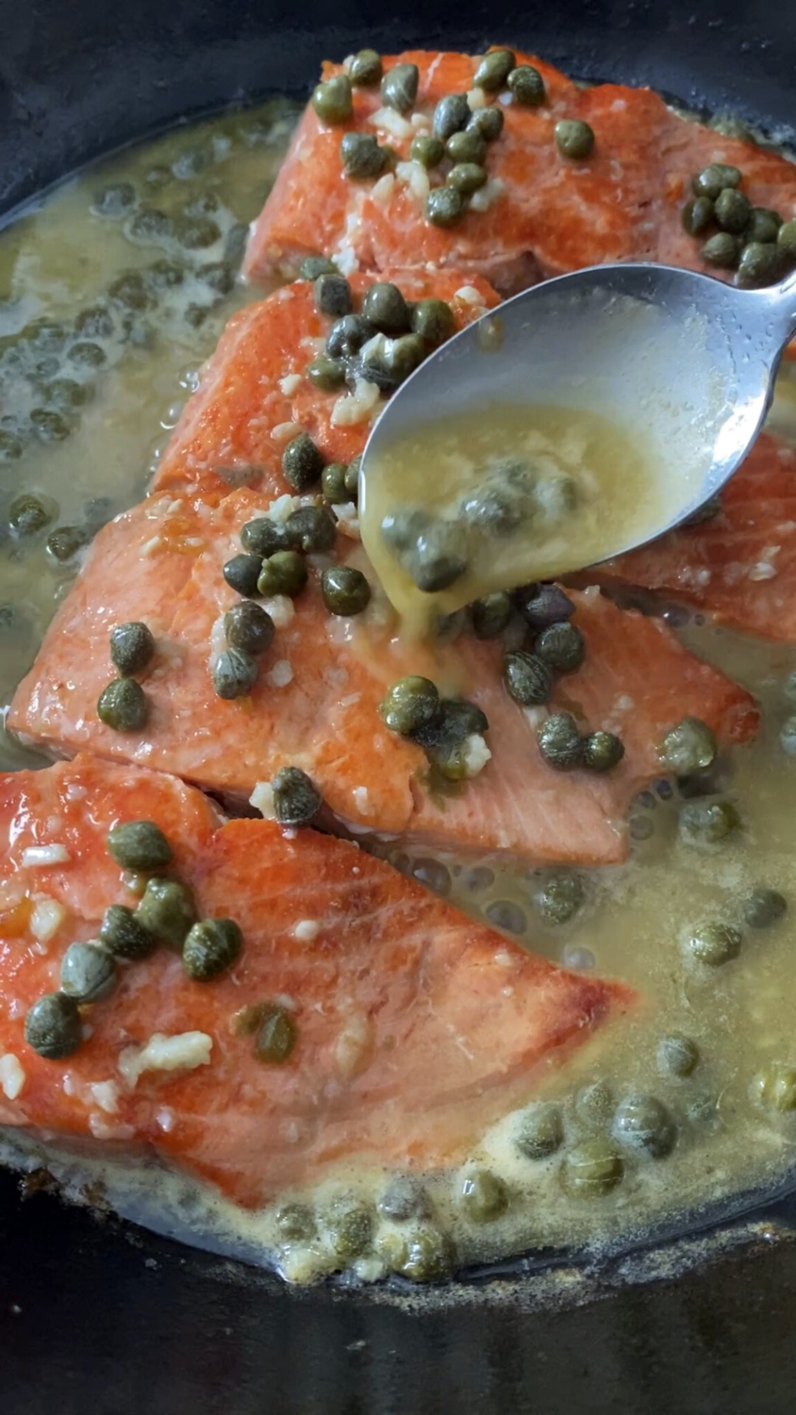 salmon piccata with lemon and capers in cast iron skillet, spooning sauce