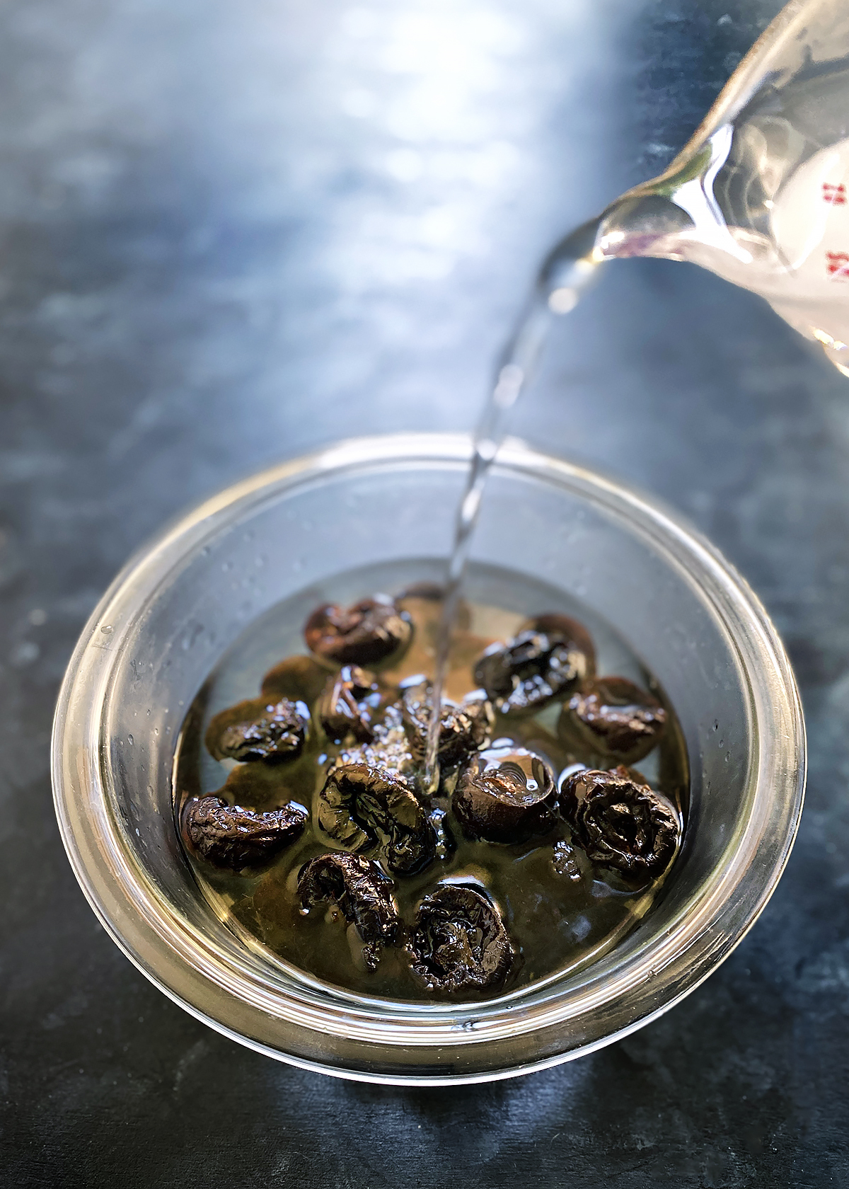 pouring water over prunes in glass mixing bowl on blue counter