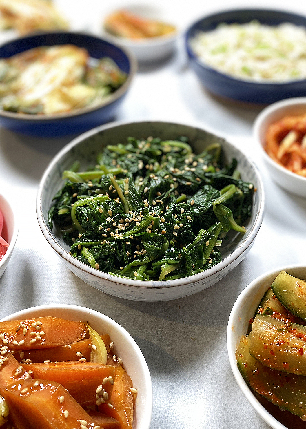 korean spinach in white bowl surrounded by assorted korean vegetable banchans: kimchi, oi kimchi, spicy marinated radish, bean sprouts, carrots, pickled daikon