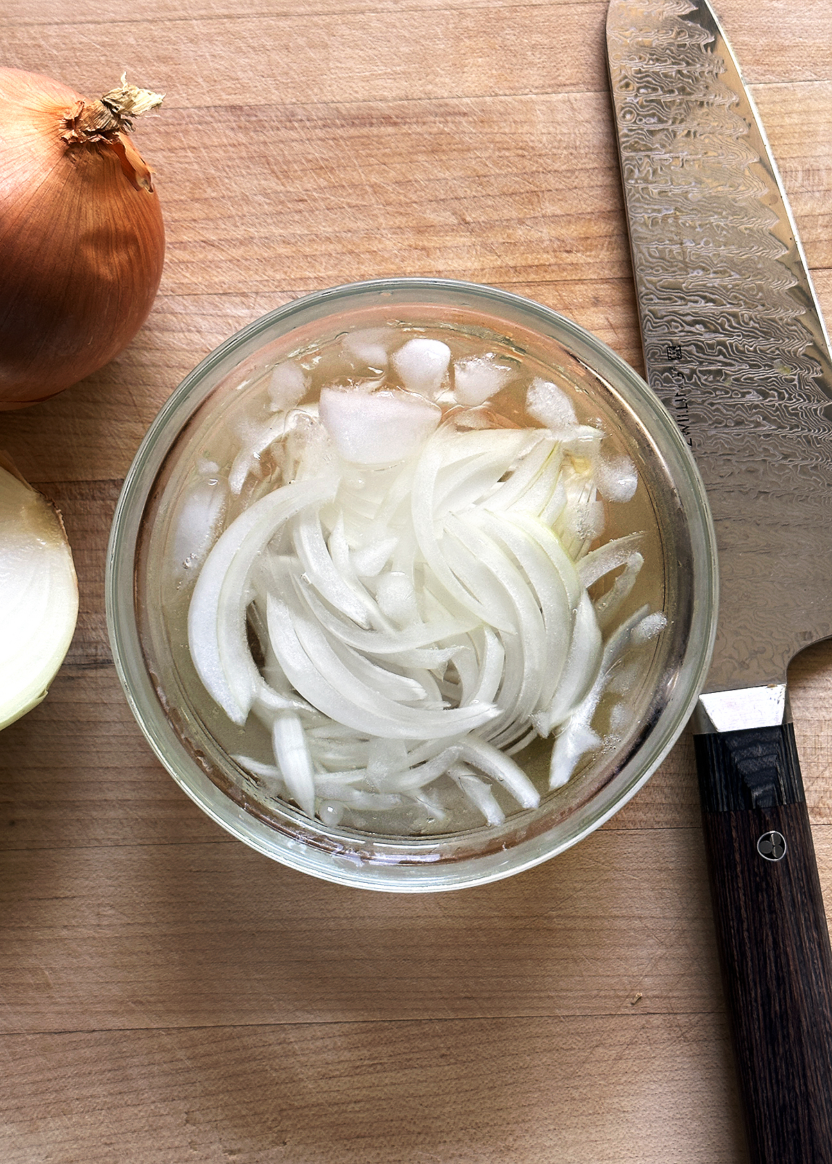 sliced onions in ice water in glass bowl on wooden cutting board