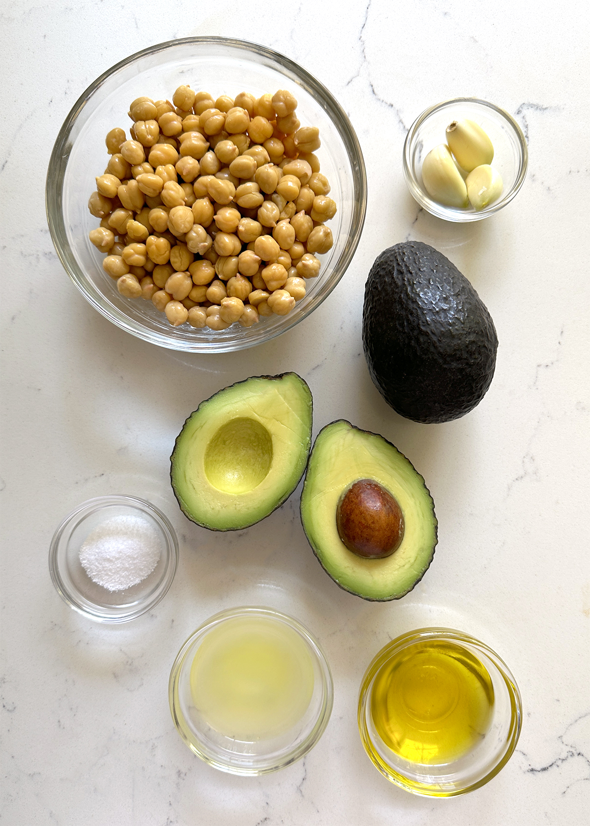 ingredients for Avocado Hummus, laid out: chickpeas, avocados cut open, peeled garlic cloves, salt lemon juice olive oil in glass bowls