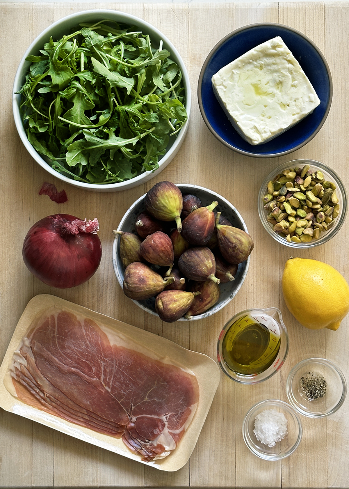 fig salad ingredients: fig, arugula, feta, prosciutto, pistachios on wooden cutting board